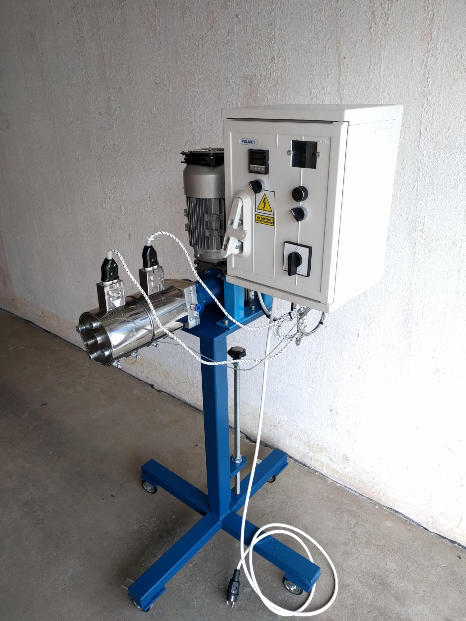 Stacjonarne urządzenie z metalowym cylindrem, silnikiem elektrycznym i panelem sterowania, osadzone na niebieskiej ramie z kółkami, widoczne kable i wtyczka.