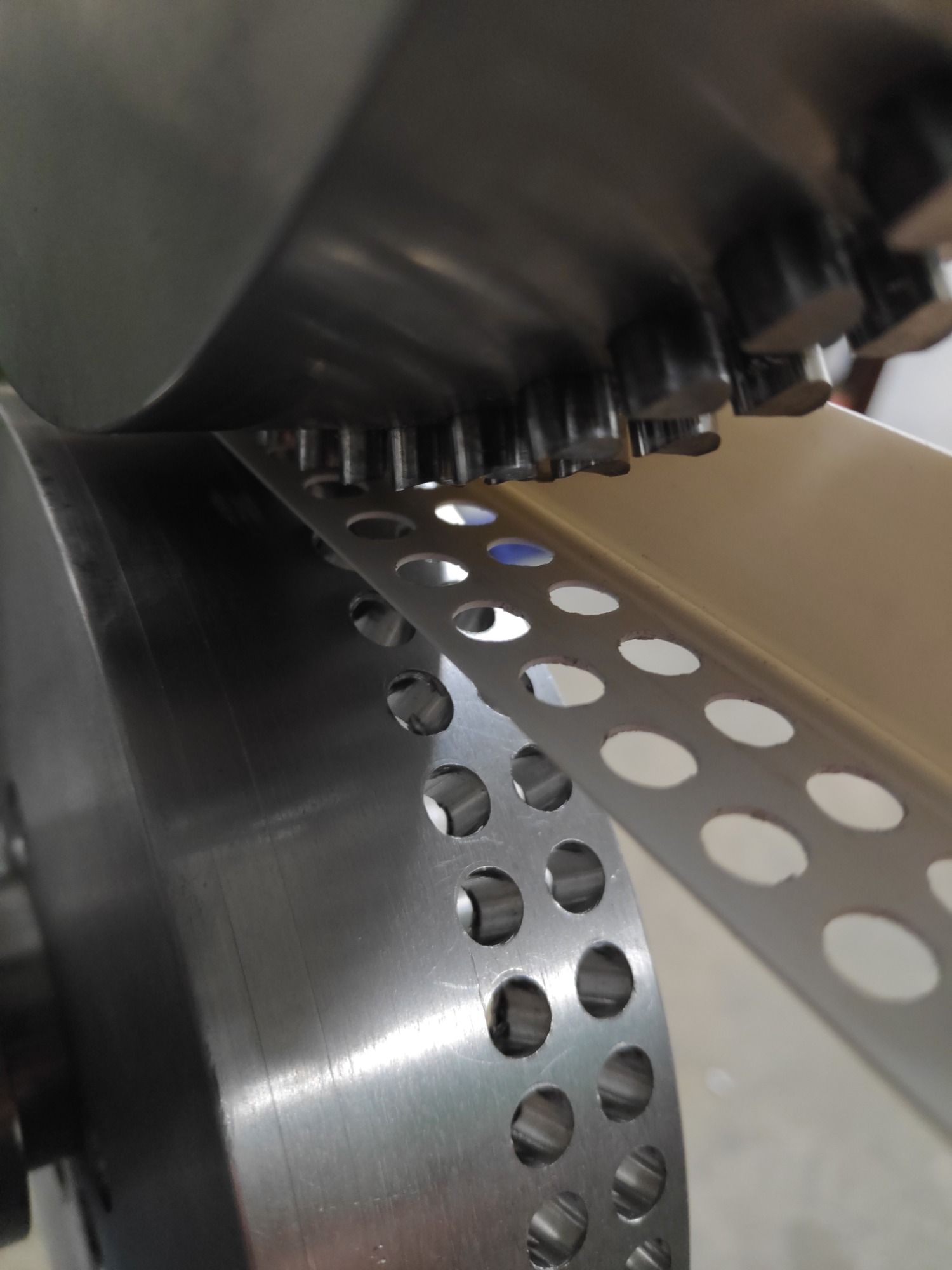 Metalowy profil z perforacją przechodzi przez maszynę z zębatymi rolkami, zbliżenie na proces dziurkowania.