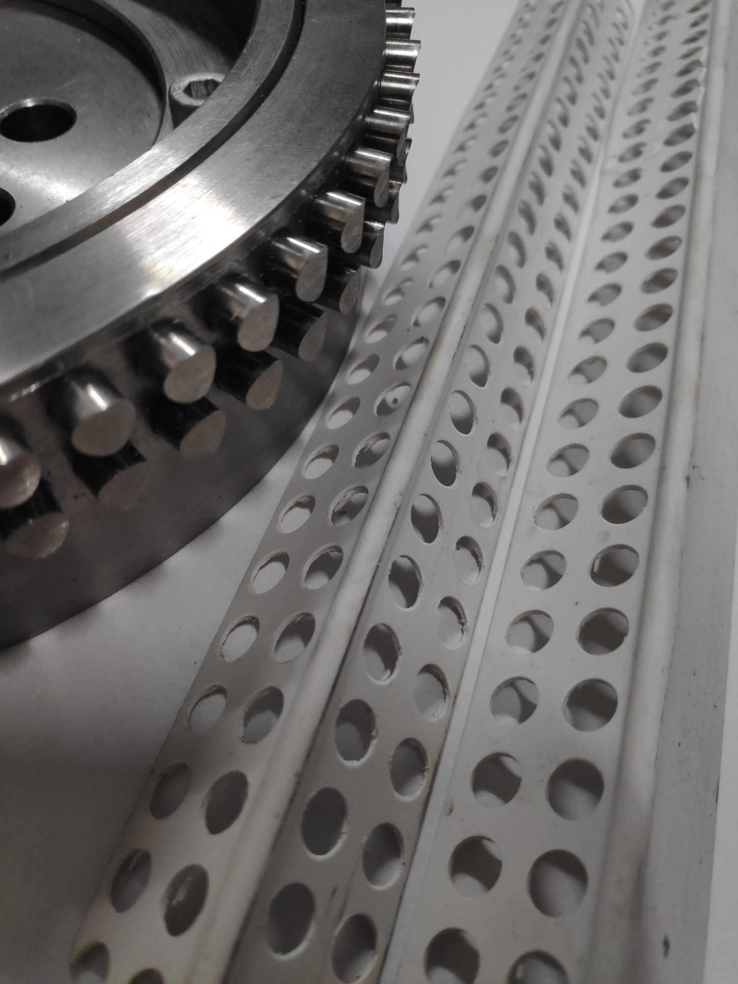 Metalowa tarcza z wypustkami oraz trzy białe, perforowane listwy z otworami ułożone na jasnym tle.