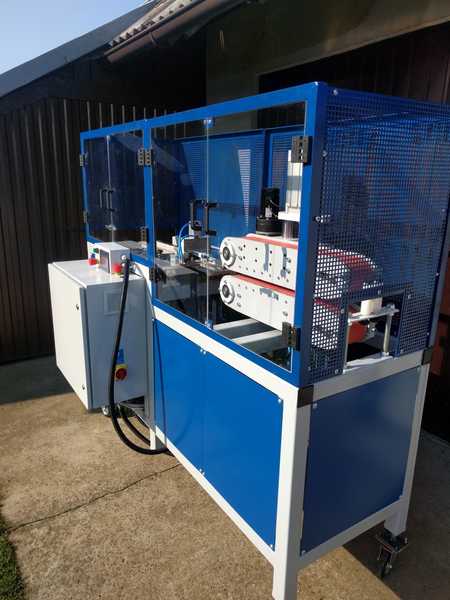 Niebiesko-biała maszyna CNC z osłoną bezpieczeństwa i panelem sterowania, widoczna na zewnątrz budynku.