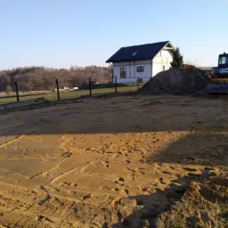 Żółty teren wyrównany pod budowę domu jednorodzinnego, z widocznym kopcem ziemi i koparką JCB w tle, ogrodzenie z siatki i drzewa w oddali.