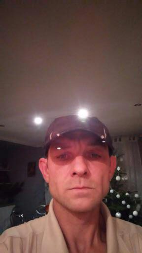 Portret mężczyzny w czapce z daszkiem, z choinką w tle i oświetleniem sufitowym. Nietypowe ujęcie z bliska, ukazujące detale twarzy i świąteczną atmosferę.