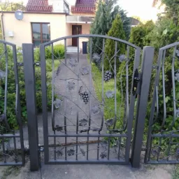 Otwarta, metalowa brama z motywem winogron prowadząca do domu z beżową elewacją i czerwonym dachem, widok z zewnątrz.