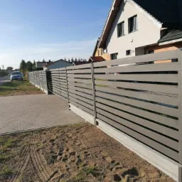 Nowoczesne ogrodzenie posesji z poziomych, szarych paneli metalowych, z widocznymi śladami opon na piasku przed ogrodzeniem i domem w tle.