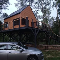 Drewniany domek letniskowy na palach z drewnianymi schodami i balustradą, obok zaparkowany srebrny samochód osobowy, w tle las.