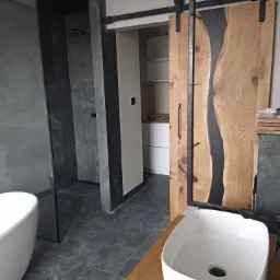 Nowoczesna łazienka z elementami drewna: przesuwna dębowa płyta drzwiowa z żywicą epoksydową, umywalka nablatowa i kabina prysznicowa z ciemnoszarego kamienia.