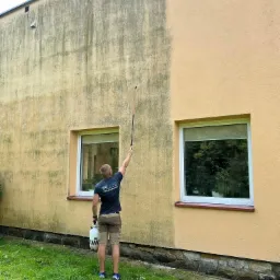 Mężczyzna w trakcie mycia elewacji budynku z żółtego tynku, widoczne wyraźne różnice w czystości powierzchni przed i po myciu, z użyciem lancy i zbiornika ciśnieniowego.