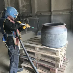 Pracownik w kombinezonie ochronnym i hełmie, z żółtymi rękawicami, czyści metalowy zbiornik umieszczony na paletach za pomocą pistoletu do obróbki strumieniowo-ściernej w hali przemysłowej.