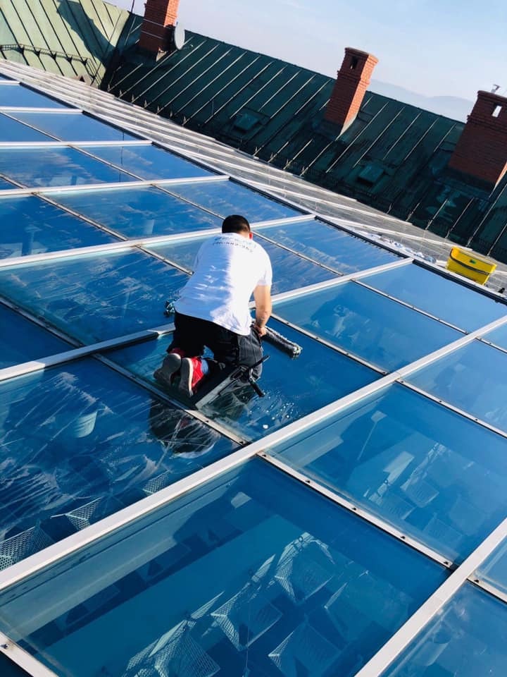 Pracownik w czerwonych butach czyści szklany dach budynku za pomocą ściągaczki, widoczny budynek w tle.
