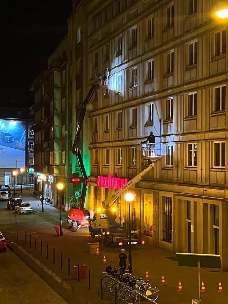 Nocne prace wysokościowe przy fasadzie kamienicy: dwóch pracowników na podnośnikach koszowych, jeden przy oświetlonym oknie, drugi wyżej, widok z dołu na ulicę z zaparkowanymi autami i rowerami.