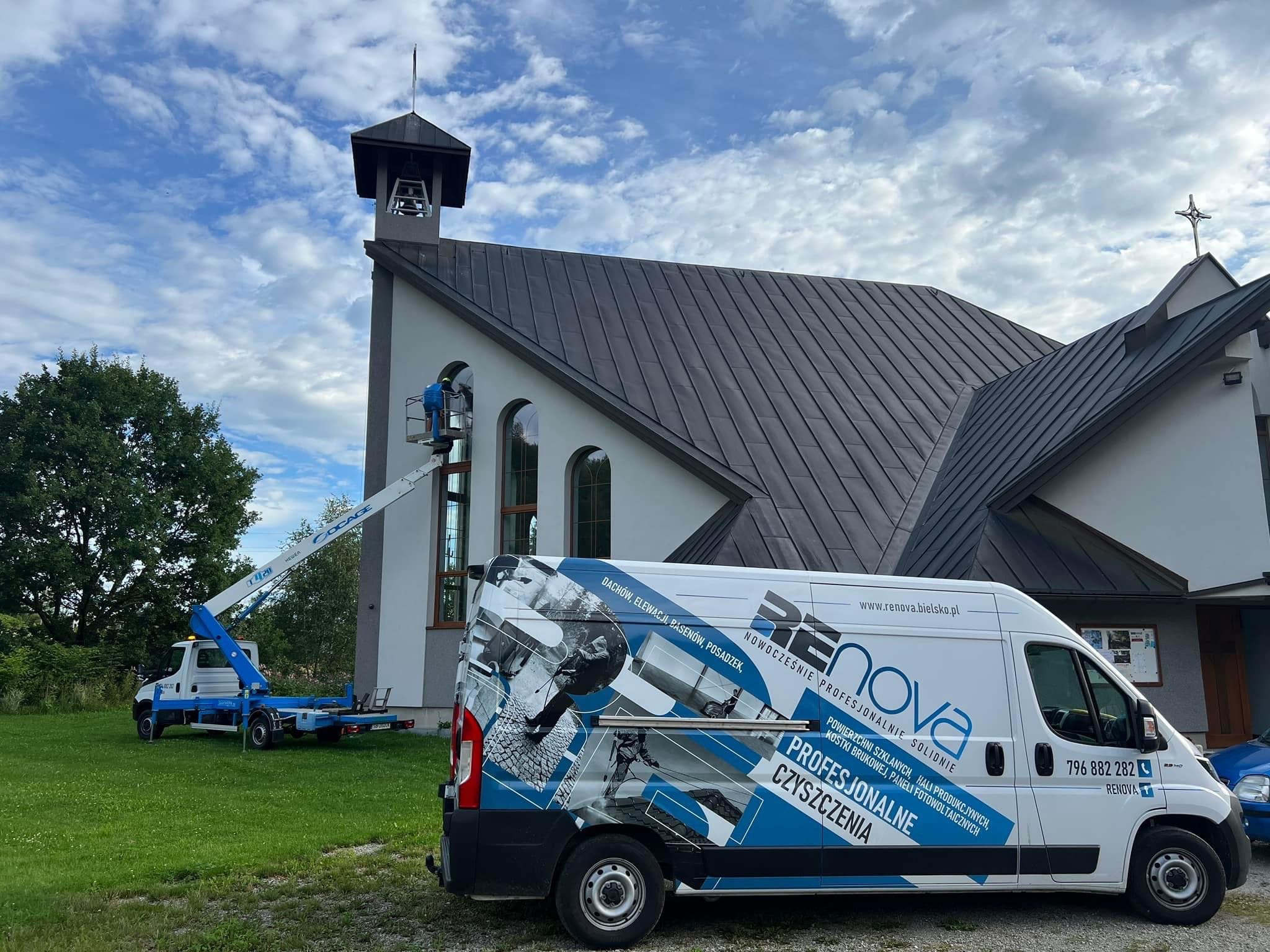 Pracownik na podnośniku koszowym czyści okna kościoła; na pierwszym planie samochód firmowy z reklamą usług czyszczenia powierzchni szklanych i elewacji.