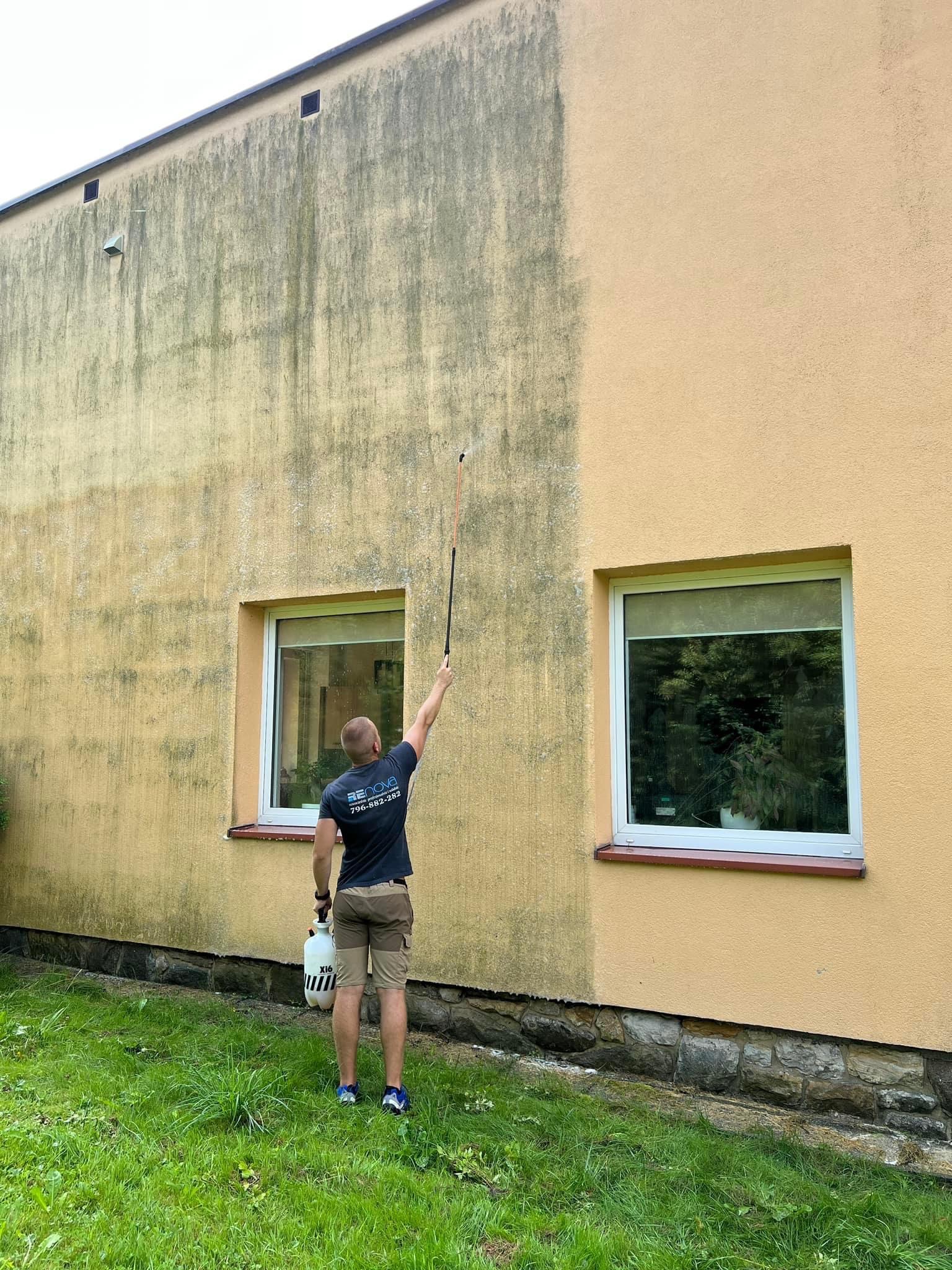 Mężczyzna w trakcie mycia elewacji budynku z żółtego tynku, widoczne wyraźne różnice w czystości powierzchni przed i po myciu, z użyciem lancy i zbiornika ciśnieniowego.
