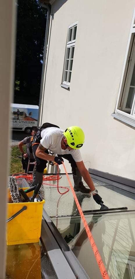 Pracownik w kasku i uprzęży alpinistycznej myje szklany dach budynku za pomocą ściągaczki, obok stoi żółte wiadro z narzędziami, w tle widoczny samochód dostawczy firmy czyszczącej.