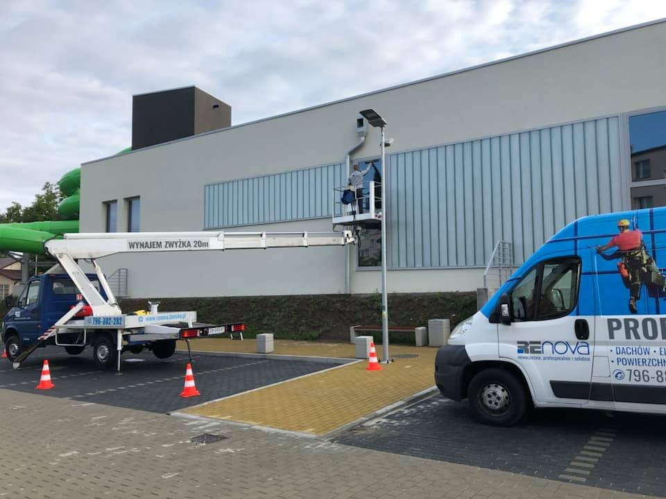 Pracownik na podnośniku koszowym czyści okna budynku użyteczności publicznej, zaparkowany samochód firmowy z logo Renova w tle, pomarańczowe pachołki bezpieczeństwa.