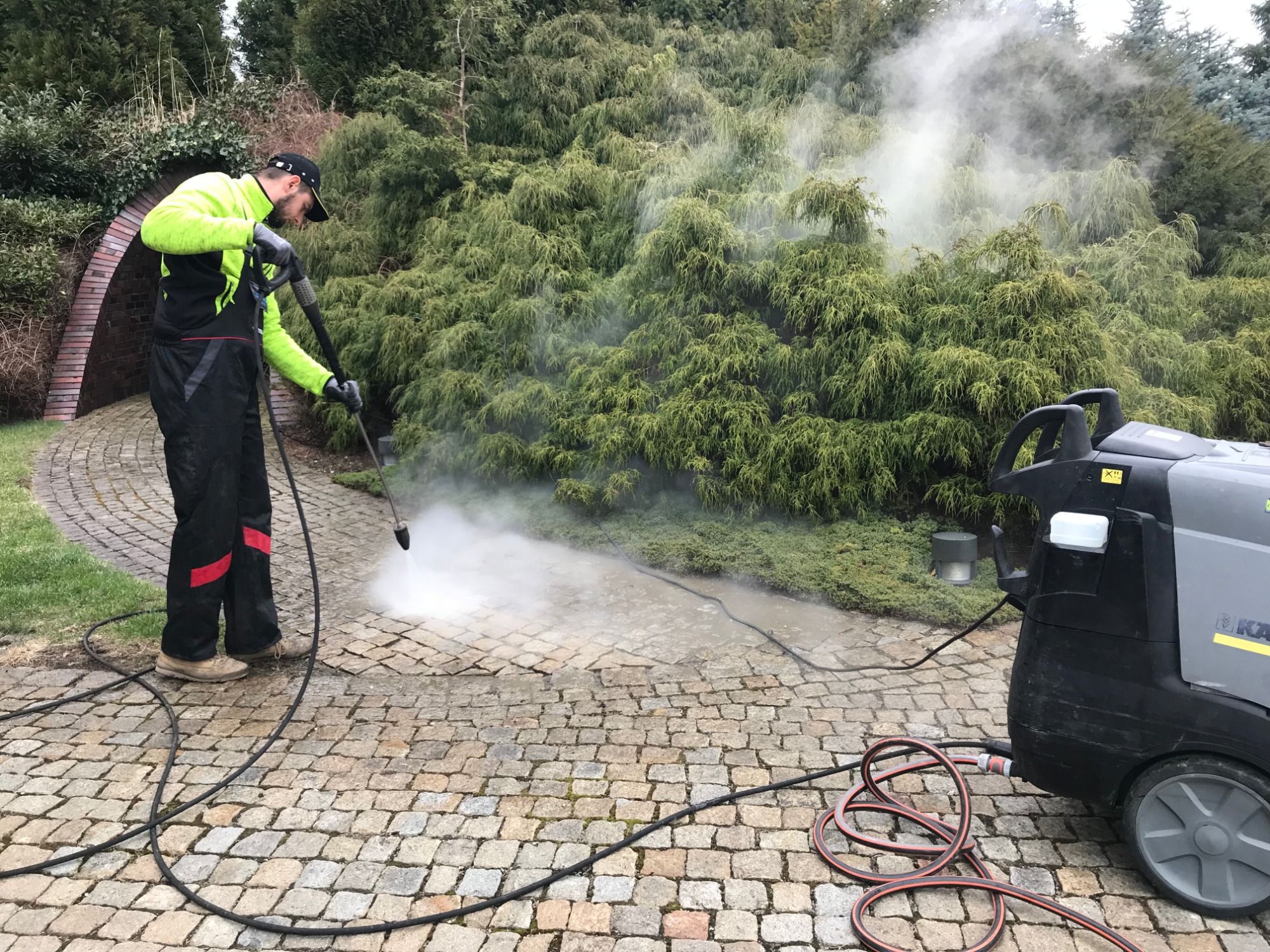 Pracownik w odzieży roboczej, z myjką ciśnieniową Kärcher, czyści kostkę brukową na podjeździe, z zielonymi krzewami w tle i łukiem z cegły.