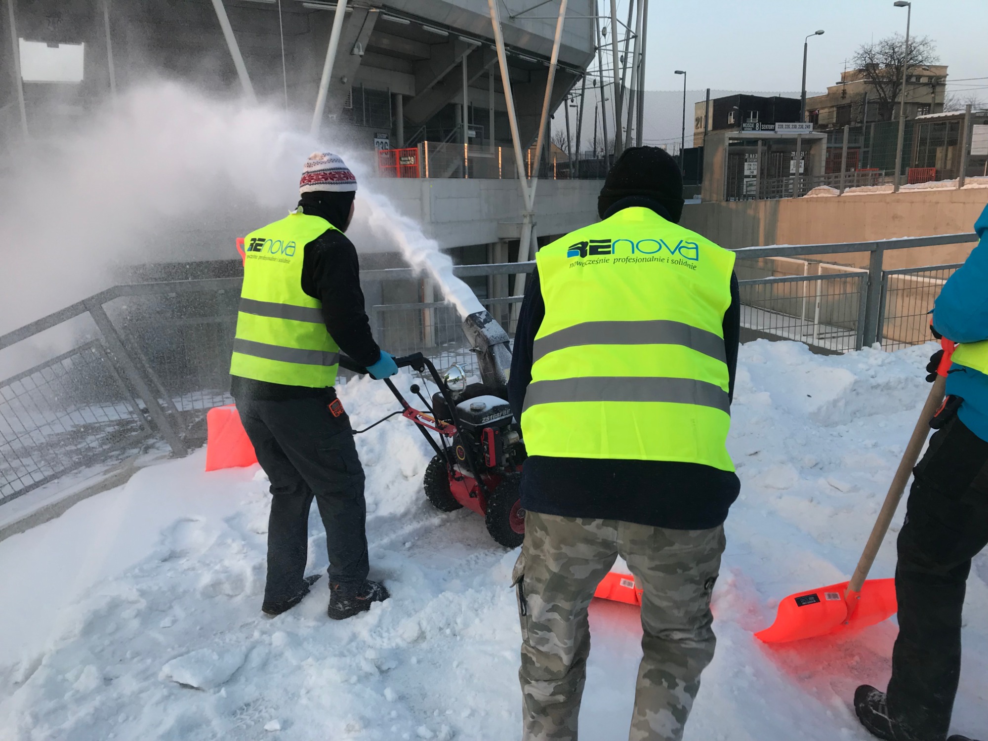 Pracownicy w odblaskowych kamizelkach z logo firmy Renova odśnieżają teren przy stadionie za pomocą odśnieżarki i łopat.