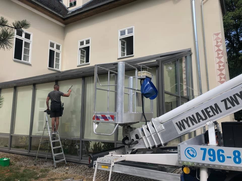 Pracownik na drabinie i podnośniku koszowym czyści okna budynku o kremowej elewacji, używając ściągaczki. Widoczne duże okna z ciemnymi ramami i ozdobny detal na ścianie.