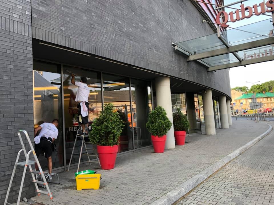 Dwóch mężczyzn w białych koszulkach czyści okna budynku Qubus Hotel, używając drabiny i ściągaczki, z widocznym żółtym pojemnikiem na narzędzia.