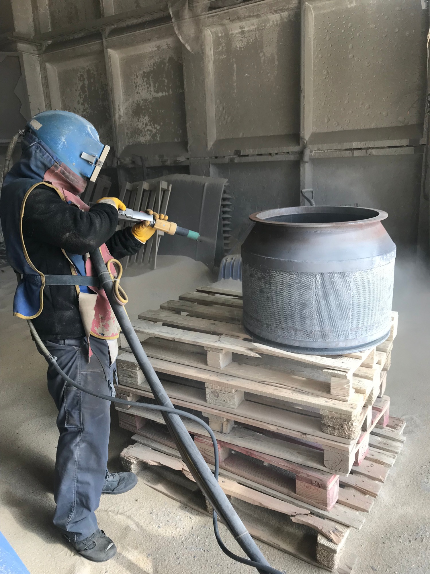 Pracownik w kombinezonie ochronnym i hełmie, z żółtymi rękawicami, czyści metalowy zbiornik umieszczony na paletach za pomocą pistoletu do obróbki strumieniowo-ściernej w hali przemysłowej.