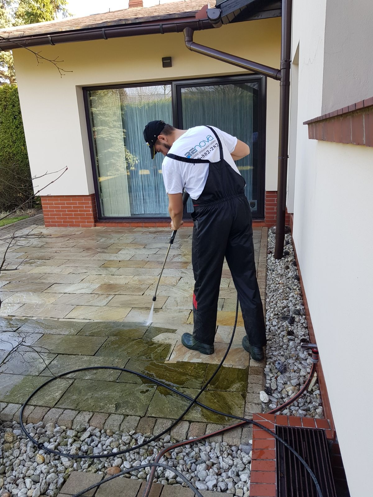 Pracownik w czarnym kombinezonie i gumowych butach czyści kostkę brukową na tarasie za pomocą myjki ciśnieniowej; widoczny efekt czyszczenia i logo firmy na koszulce.