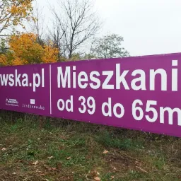 Fioletowy baner reklamowy z białym napisem 'jordanowska.pl Mieszkania od 39 do 65m2', umieszczony na drewnianych słupkach na tle jesiennego krajobrazu.