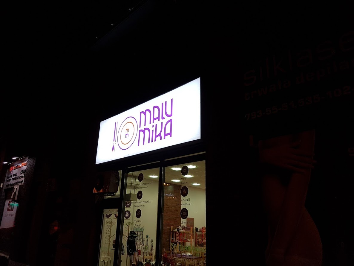 Podświetlany kaseton reklamowy z fioletowym logo 'MALU MIKA' nad wejściem do sklepu, widoczny w nocy. W tle szyld salonu kosmetycznego.