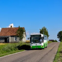 Jelcz 120MM/2 w zielono-białych barwach linii 106 wjeżdża na wzniesienie asfaltowej drogi, w tle niewielki dom z ceglanym dachem i błękitne niebo.