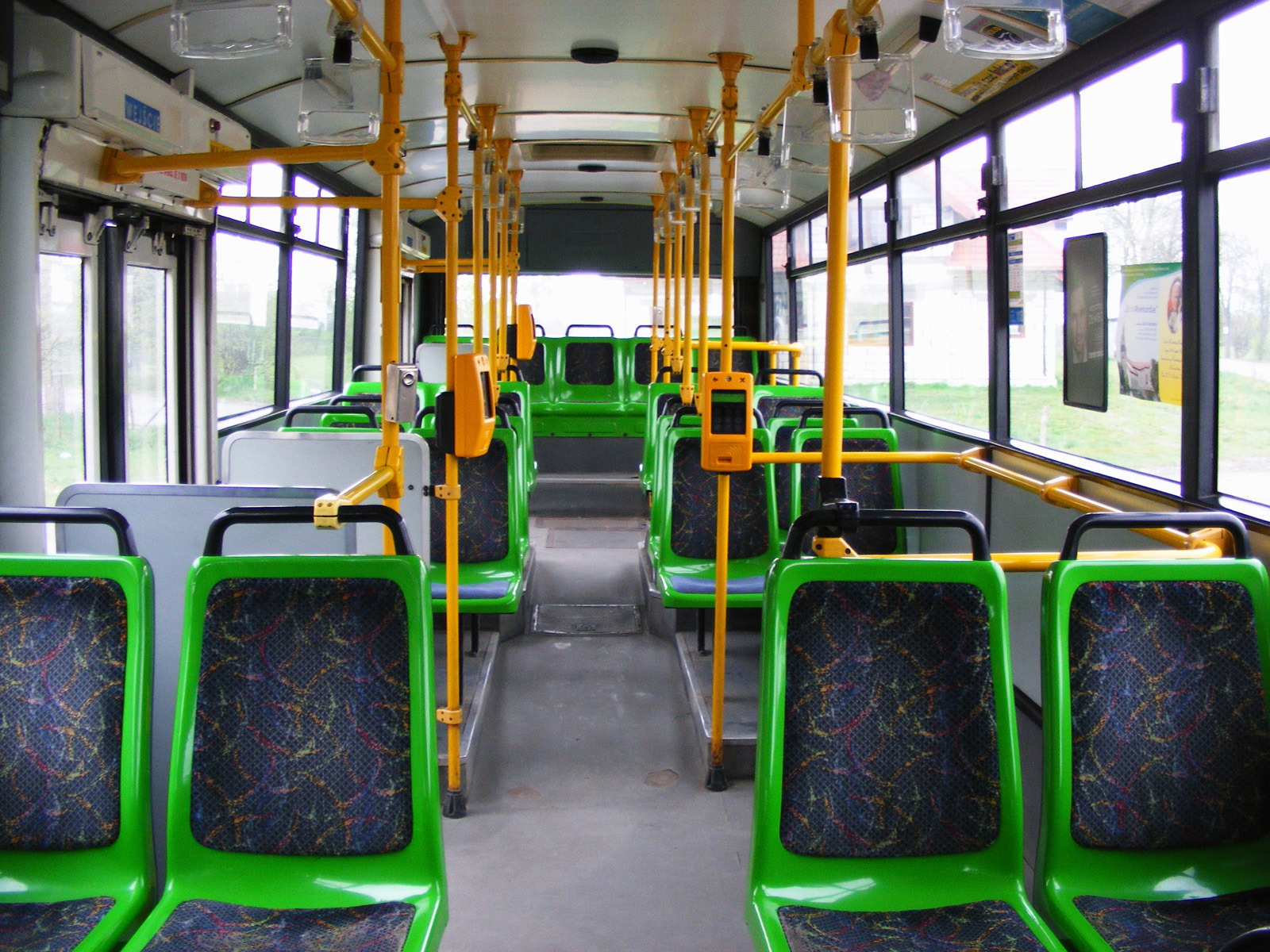 Wnętrze autobusu miejskiego z zielonymi siedzeniami i żółtymi poręczami, widok z przodu pojazdu w kierunku tylnych drzwi.