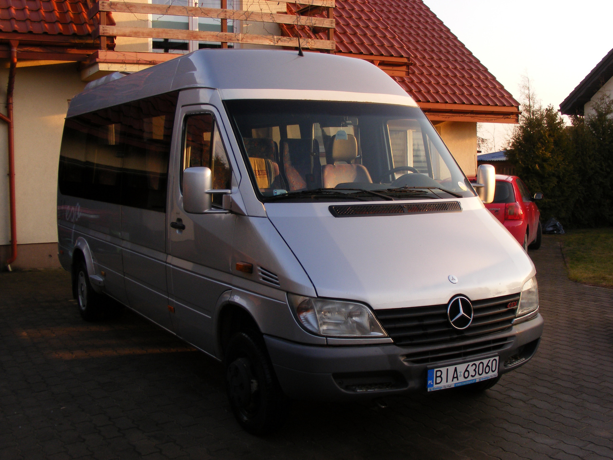 Srebrny Mercedes-Benz Sprinter 413 CDI zaparkowany na brukowanej nawierzchni przed domem z czerwoną dachówką, widoczny z przodu i boku.