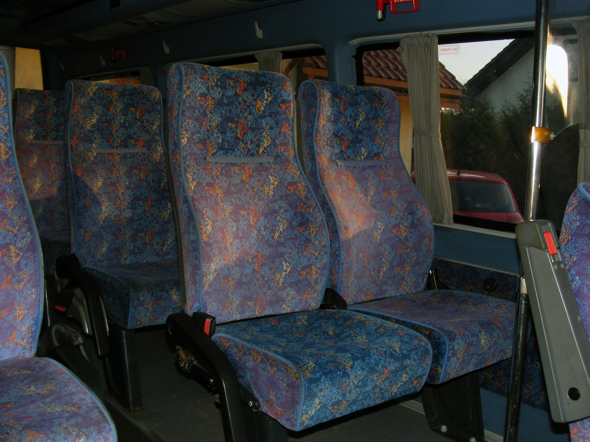 Wnętrze autobusu z rzędami niebieskich siedzeń z wzorzystą tapicerką, widok przez okno na dom i czerwony samochód.