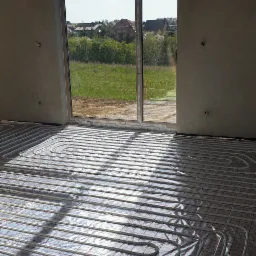 Białe rurki ogrzewania podłogowego ułożone na folii z czerwoną siatką w surowym wnętrzu domu z dużym oknem wychodzącym na zielony krajobraz.