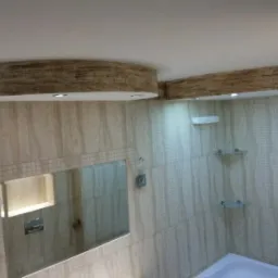 Wykończona łazienka z beżowymi płytkami imitującymi marmur, dekoracyjnymi belkami sufitowymi z efektem drewna, podświetlanym lustrem i szklanymi półkami.