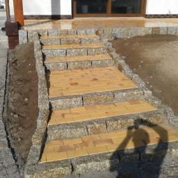 Lech-Bruk Lech Mędykowski - Żółto-brązowe schody z kostki prowadzące do drzwi wejściowych, obramowane szarym granitem, częściowo w cieniu osoby fotografującej.