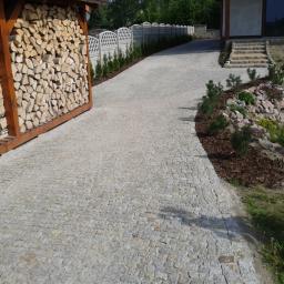 Lech-Bruk Lech Mędykowski - Podjazd z jasnoszarej kostki granitowej prowadzący do domu, obok drewniana wiata na drewno i rabata z kamieniami i roślinami.