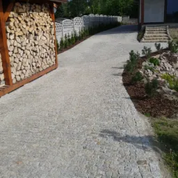 Podjazd z jasnoszarej kostki granitowej prowadzący do domu, obok drewniana wiata na drewno i rabata z kamieniami i roślinami.