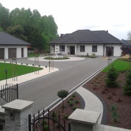 Lech-Bruk Lech Mędykowski - Elegancki podjazd do domu z fontanną na rondzie, wykończony szarą kostką i białym żwirem, z symetrycznie posadzonymi drzewkami i krzewami.