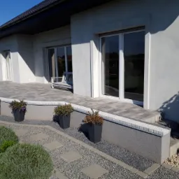 Nowoczesny taras z kostki brukowej w odcieniach szarości, z niskim murkiem obłożonym dekoracyjną kostką, donice z kwiatami i ścieżka z kamieni na żwirowym podłożu, na tle jasnego domu z dużymi oknami.