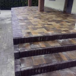 Lech-Bruk Lech Mędykowski - Wykończone schody zewnętrzne z kostki brukowej w odcieniach brązu i beżu, prowadzące na taras z kostki, z widocznymi kolumnami i fragmentem samochodu dostawczego w tle.