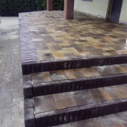 Wykończone schody zewnętrzne z kostki brukowej w odcieniach brązu i beżu, prowadzące na taras z kostki, z widocznymi kolumnami i fragmentem samochodu dostawczego w tle.