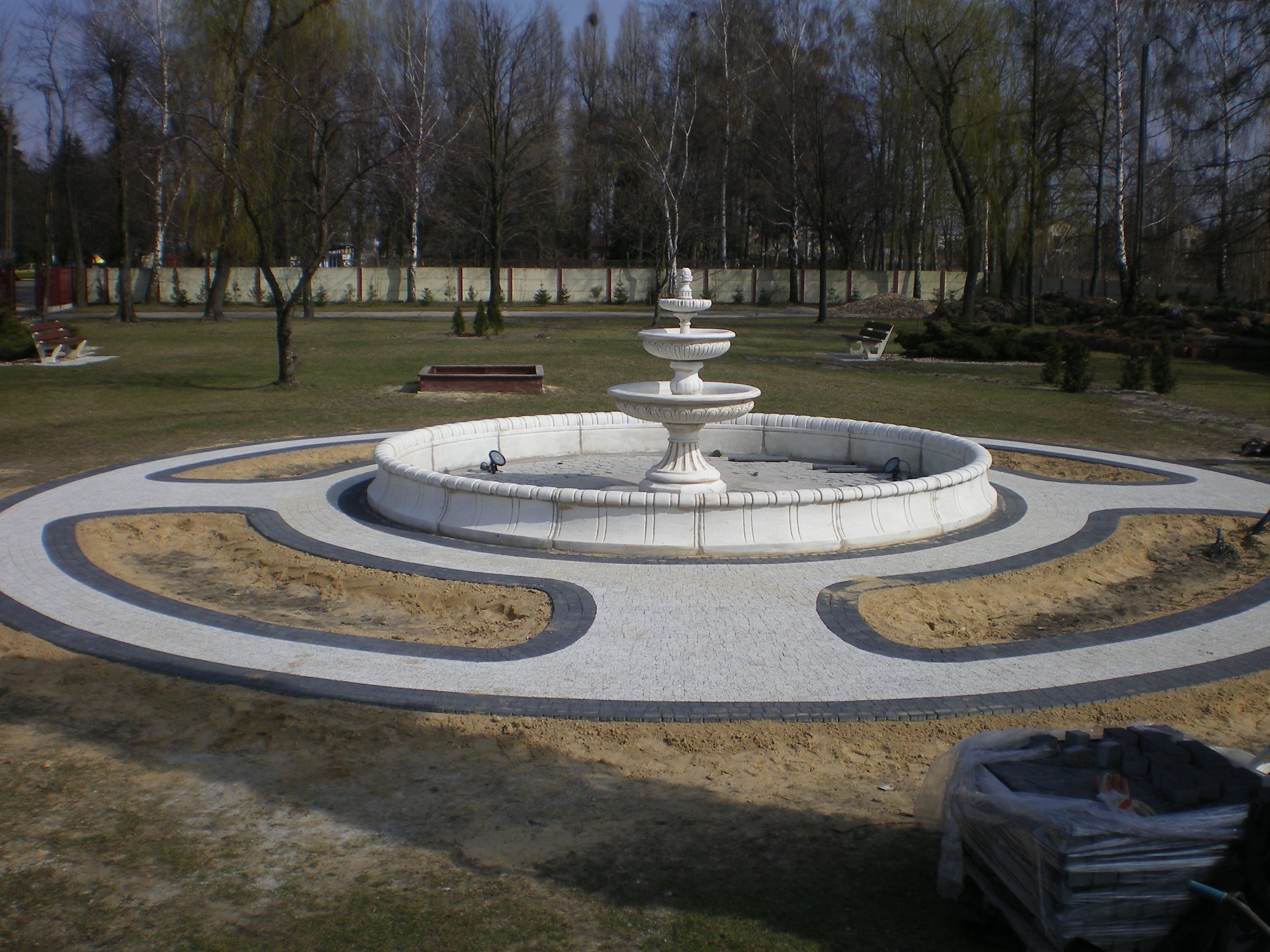 Okrągła fontanna otoczona ścieżką z kostki brukowej w parku, widoczne drzewa bez liści w tle i paleta z kostką brukową w folii na pierwszym planie.