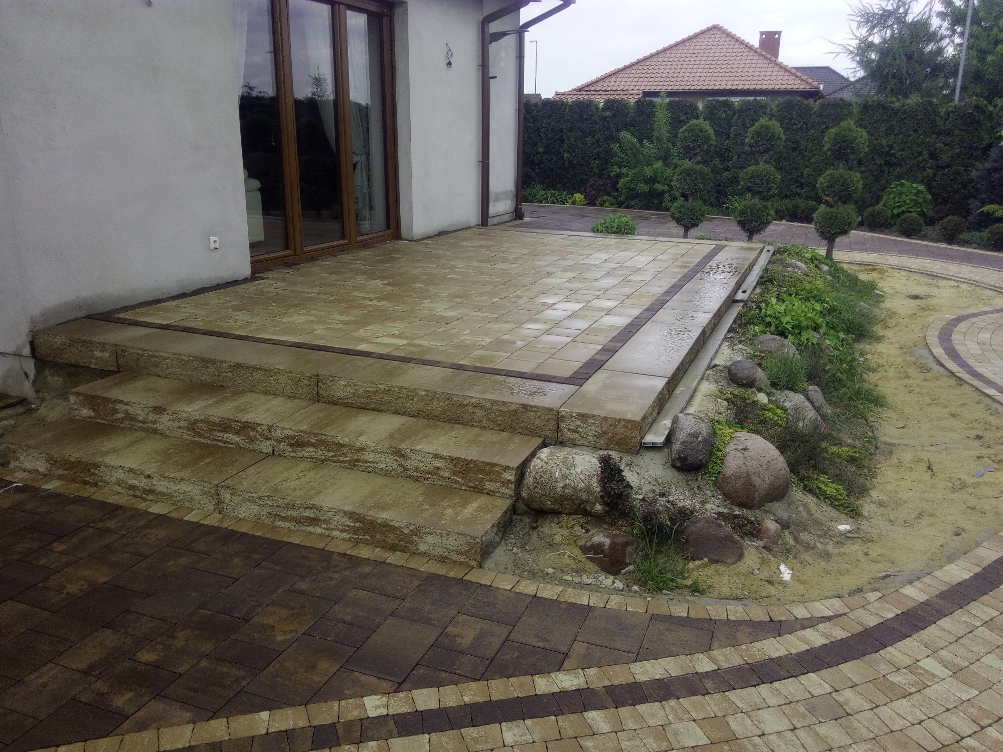 Wyłożony kostką taras z trzema stopniami, obrzeżem z ciemniejszej kostki i odpływem liniowym, widok na ogród z tujami i fragmentem dachu domu.
