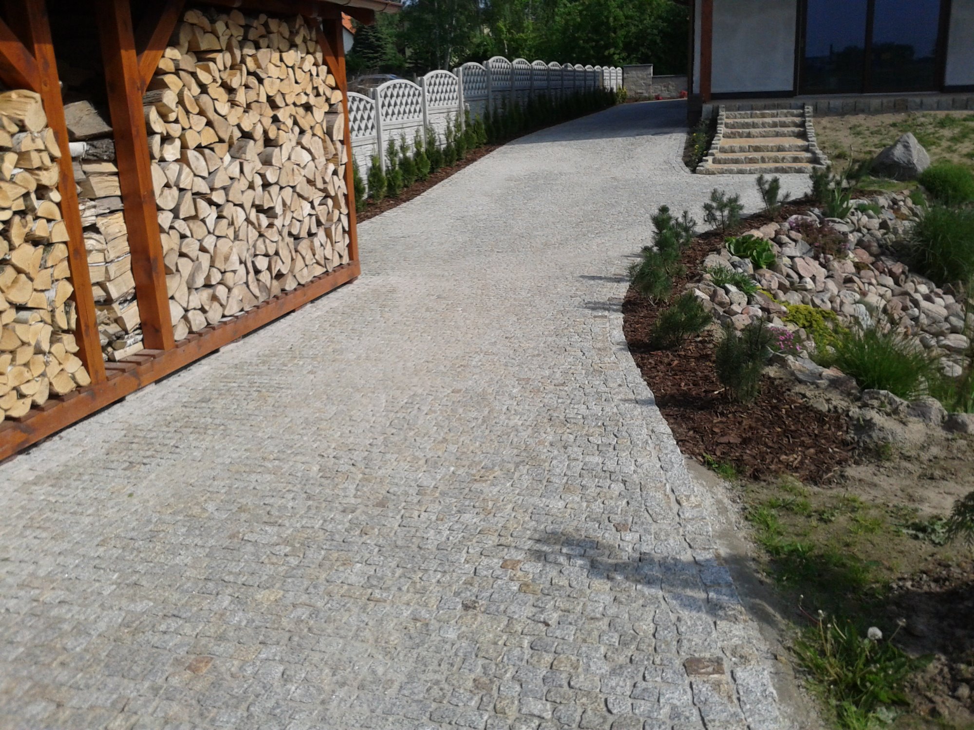 Podjazd z jasnoszarej kostki granitowej prowadzący do domu, obok drewniana wiata na drewno i rabata z kamieniami i roślinami.