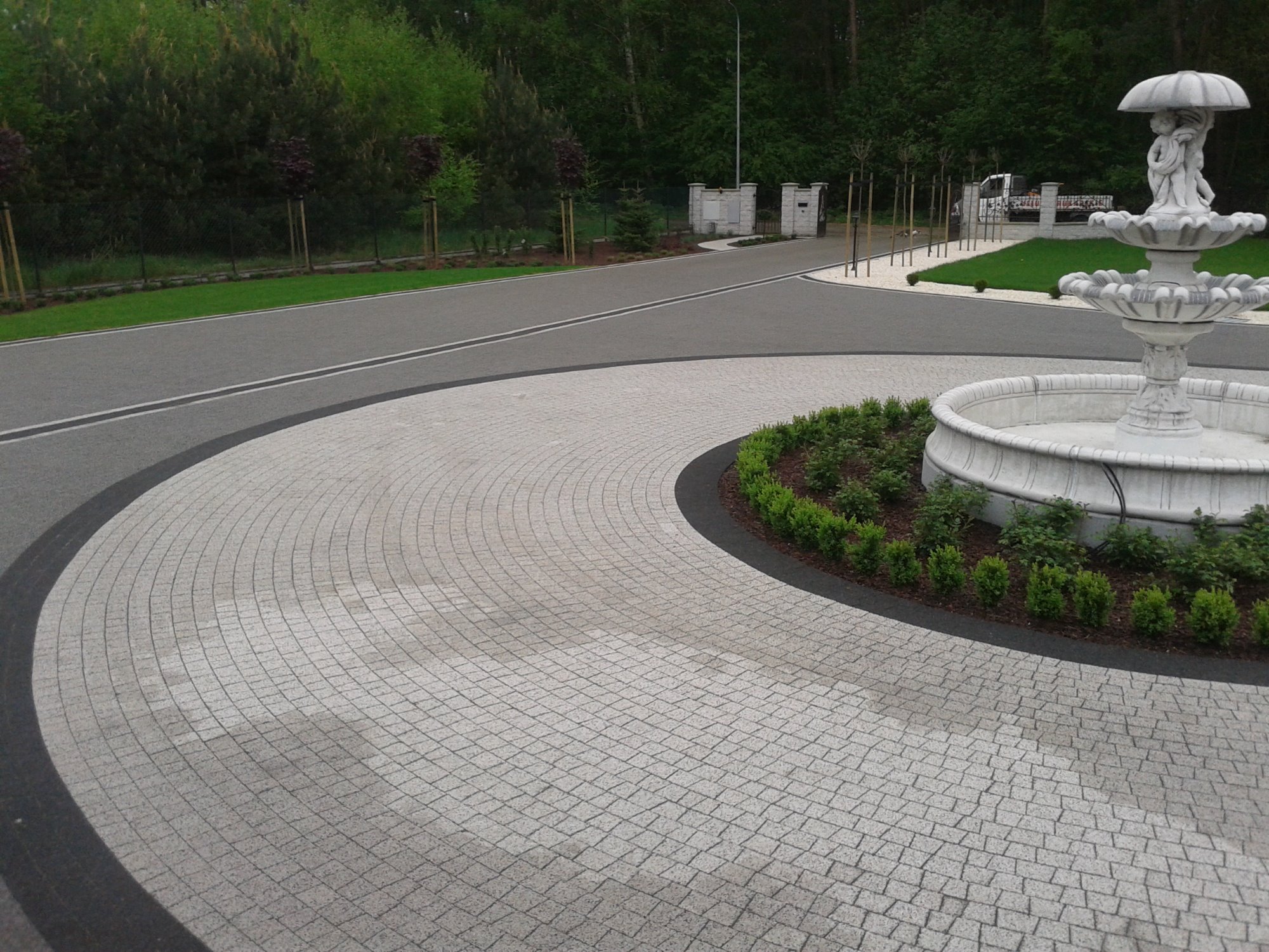 Podjazd wyłożony szarą kostką granitową w kształcie wachlarza, obrzeże z ciemnej kostki, w tle fontanna i zadbana zieleń.