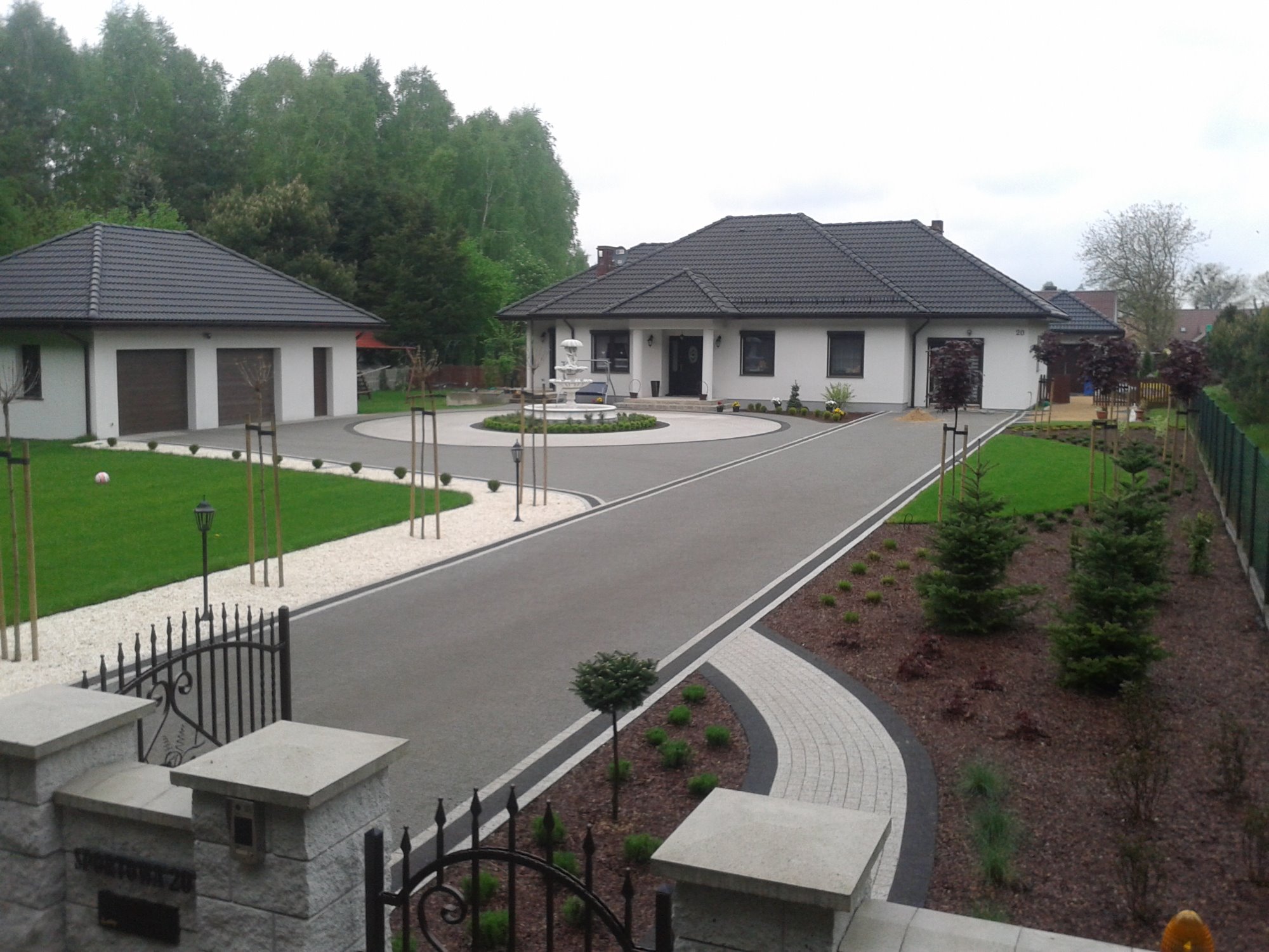Elegancki podjazd do domu z fontanną na rondzie, wykończony szarą kostką i białym żwirem, z symetrycznie posadzonymi drzewkami i krzewami.