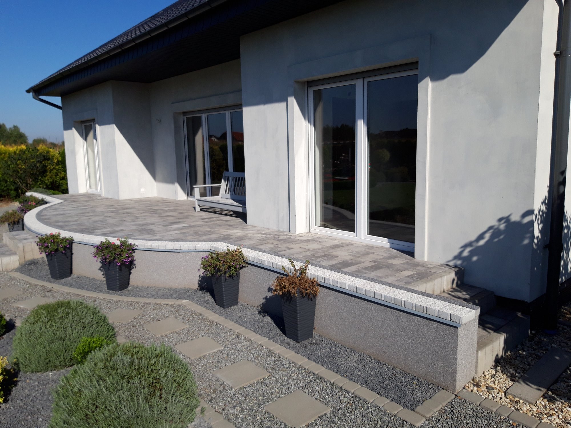 Nowoczesny taras z kostki brukowej w odcieniach szarości, z niskim murkiem obłożonym dekoracyjną kostką, donice z kwiatami i ścieżka z kamieni na żwirowym podłożu, na tle jasnego domu z dużymi oknami.
