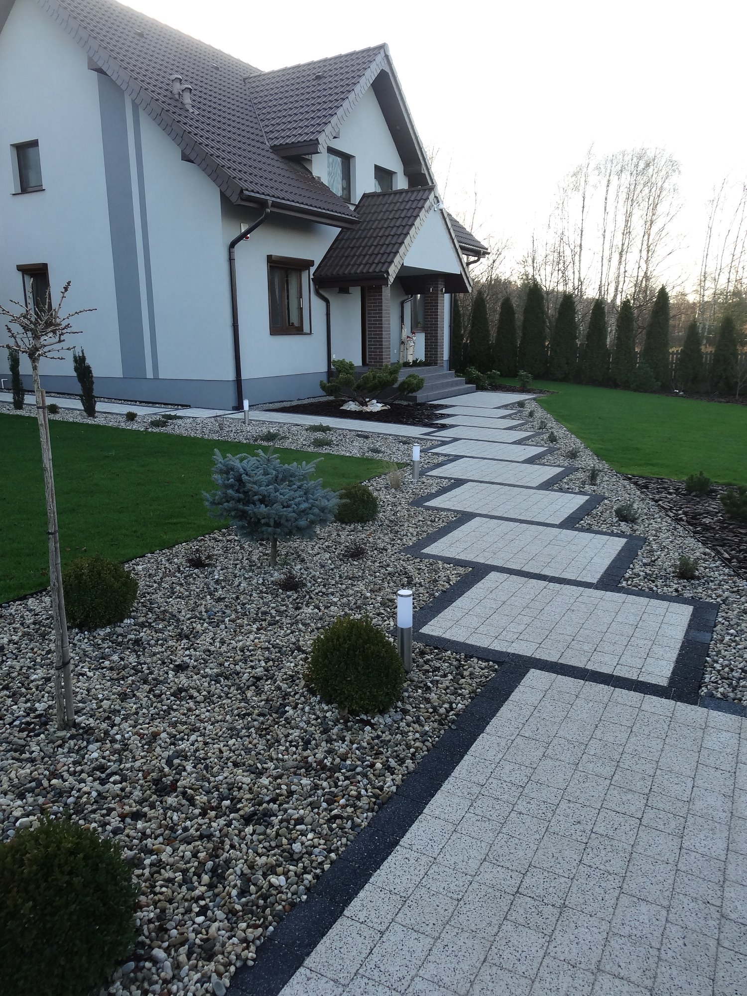 Ogród z modernistycznym domem i ścieżką z jasnoszarej kostki brukowej z ciemną obwódką, otoczoną białymi kamykami i niską zielenią.