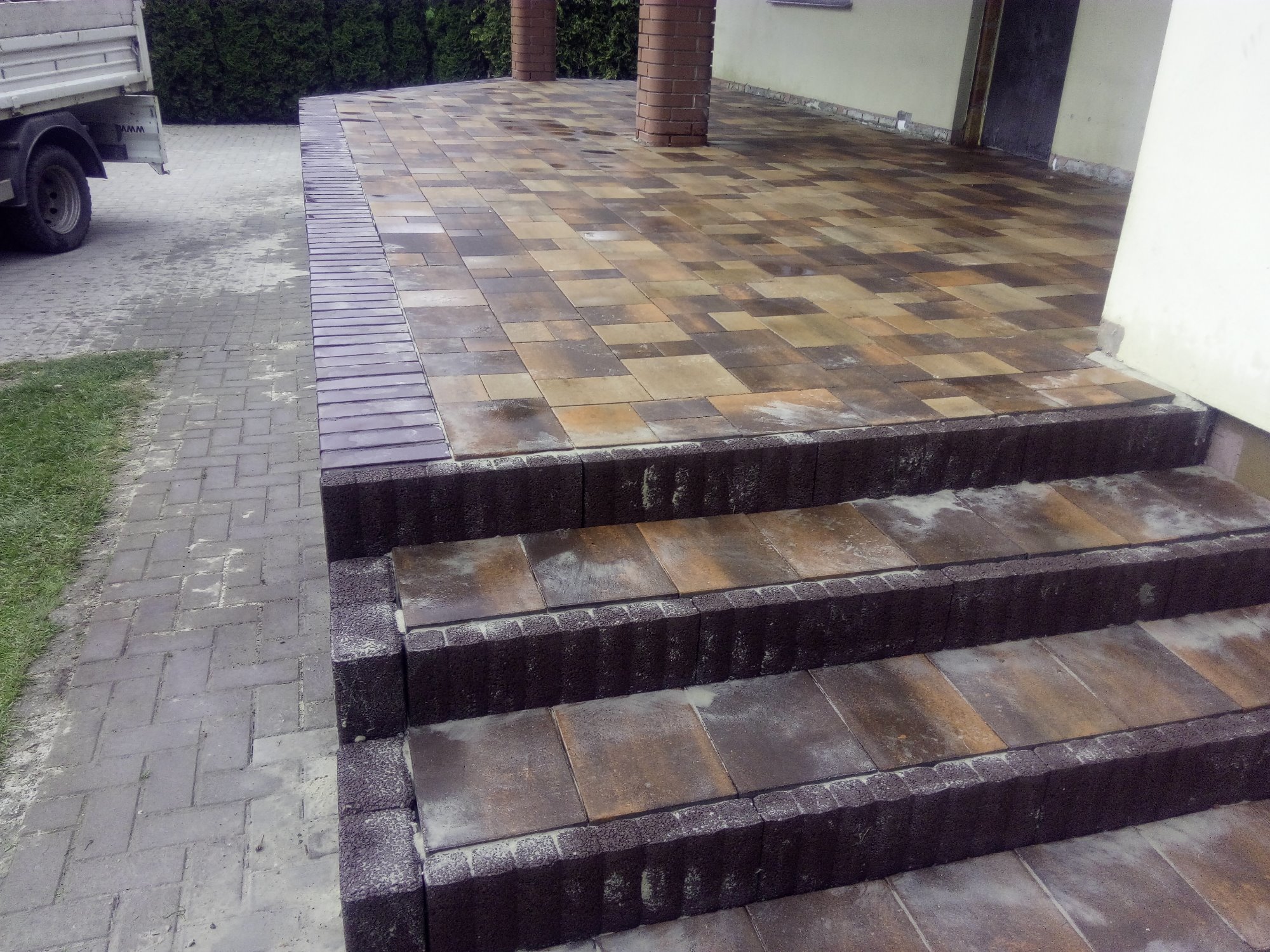 Wykończone schody zewnętrzne z kostki brukowej w odcieniach brązu i beżu, prowadzące na taras z kostki, z widocznymi kolumnami i fragmentem samochodu dostawczego w tle.