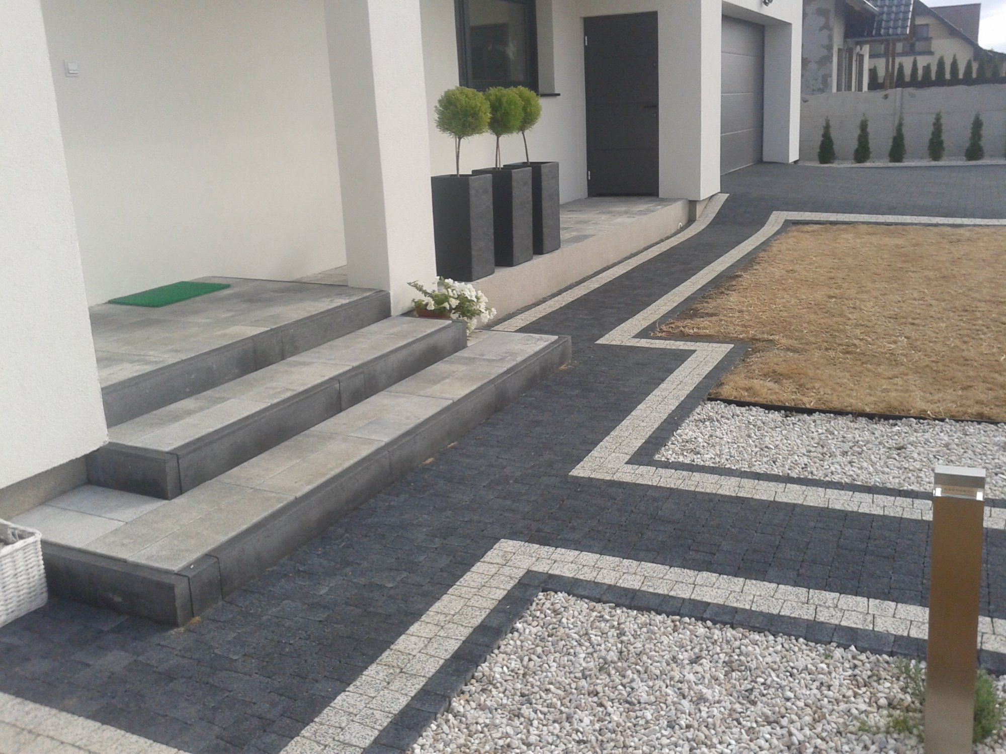 Nowoczesne wejście do domu z kamiennymi schodami, ciemną kostką brukową obramowaną jasną kostką, żwirowym wypełnieniem i trzema topiarami w wysokich donicach.