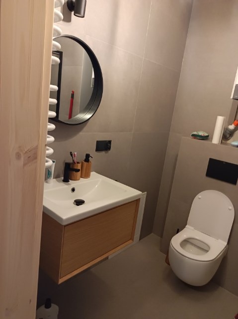 Nowoczesna łazienka z szarą glazurą, wiszącą szafką pod umywalką z drewnianym frontem, okrągłym lustrem i podwieszaną toaletą.