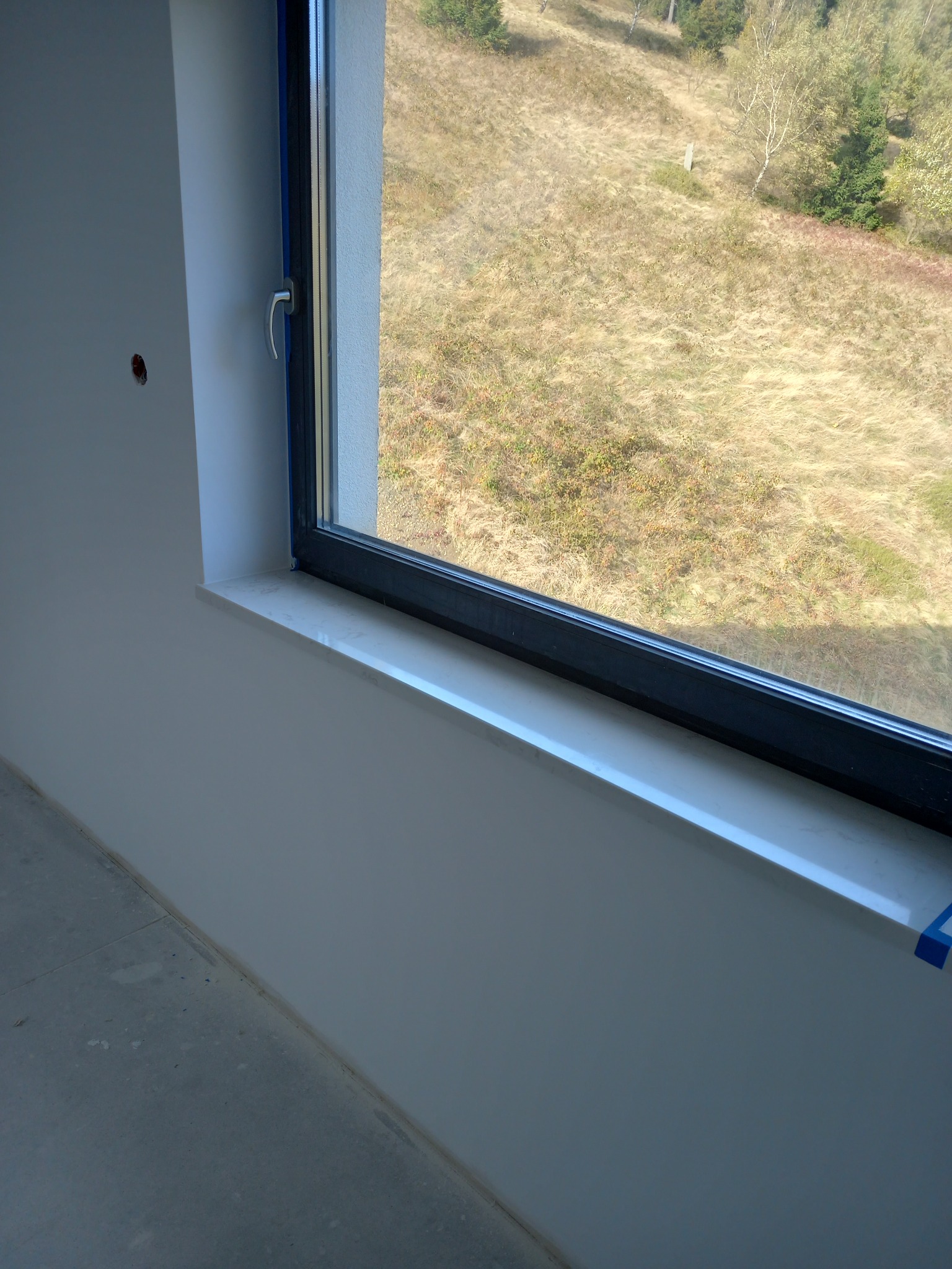 Nowe okno z jasnym parapetem, zabezpieczone niebieską taśmą malarską, widok na zewnątrz na łąkę i drzewa w słoneczny dzień.